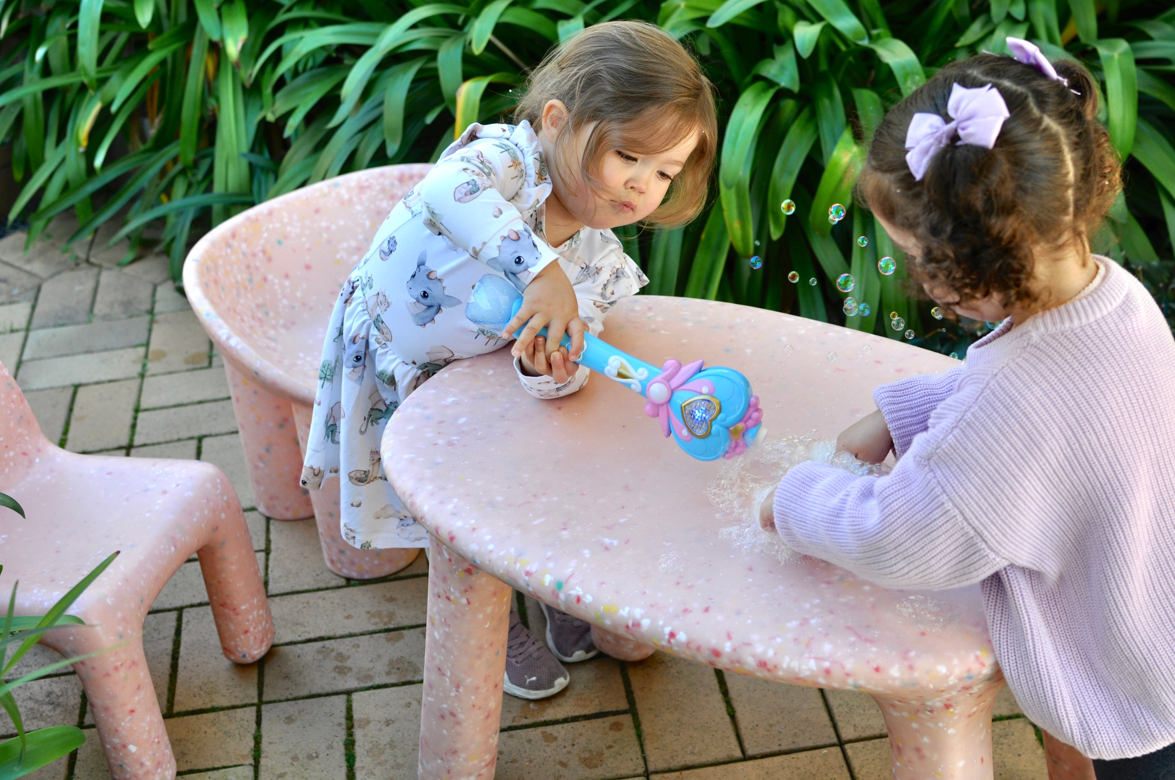 Two young children playing with bubble toys at a small pink speckles confetti table and chairs set outdoors.