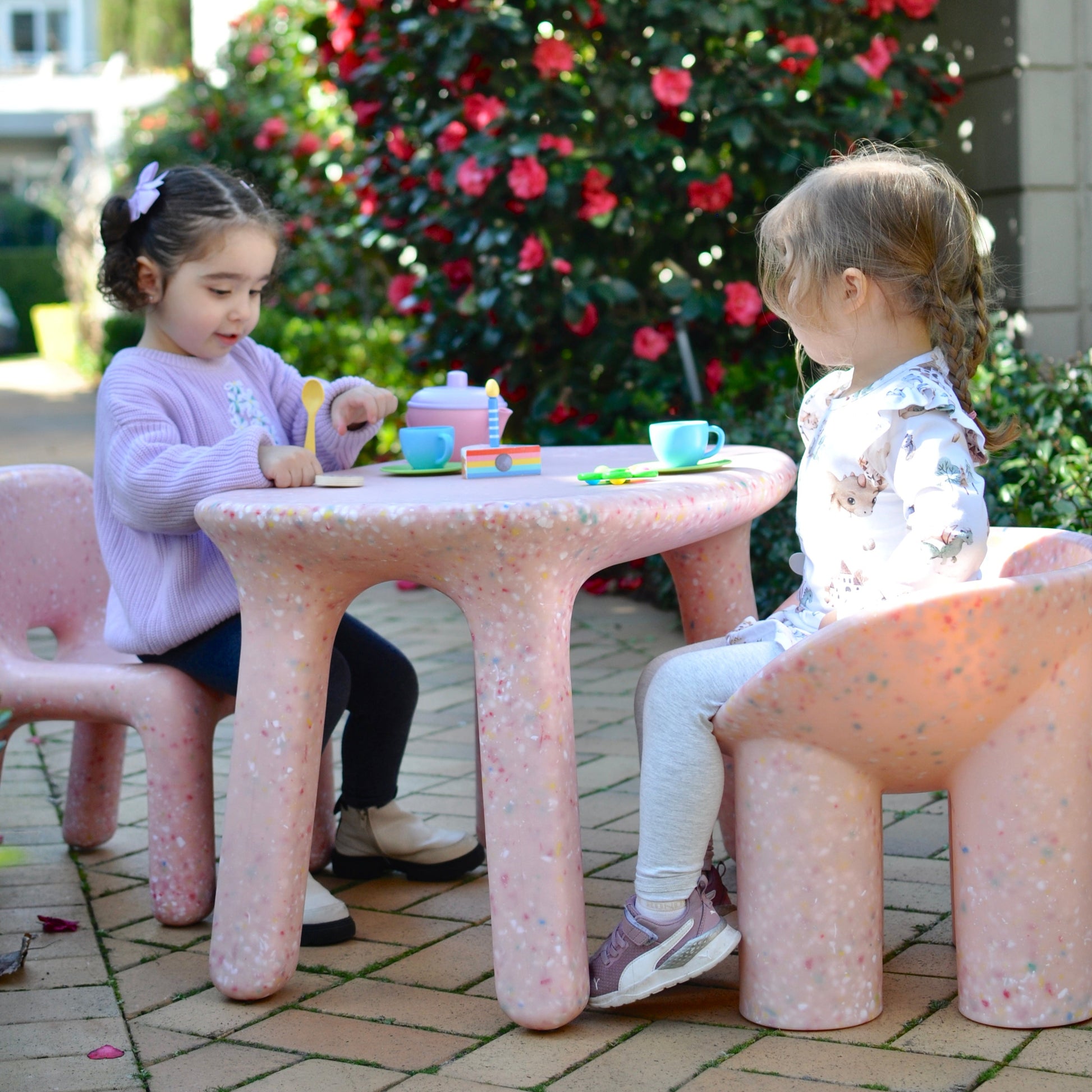 Two children playing at a pink speckles confetti table and chairs outdoors with a flowering bush in the background.
