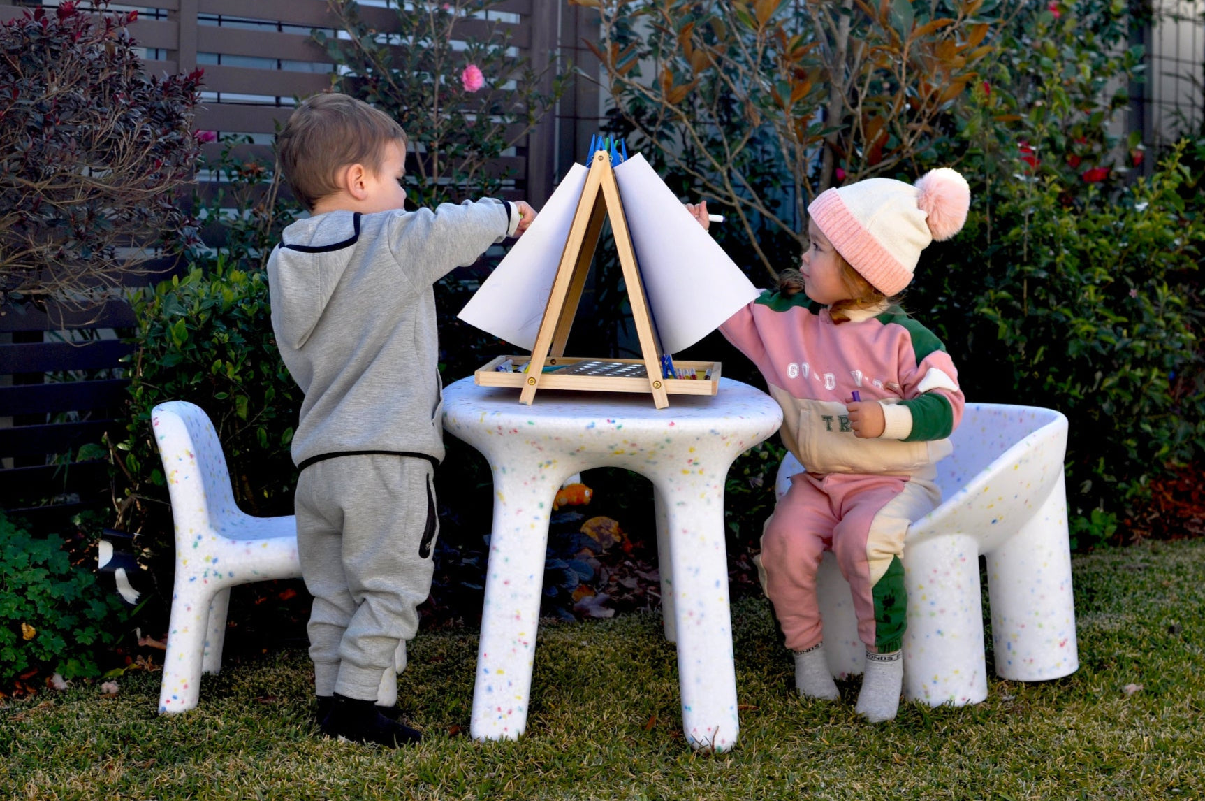 girl and boy painting in the garden, sitting on white confetti tables and chairs set