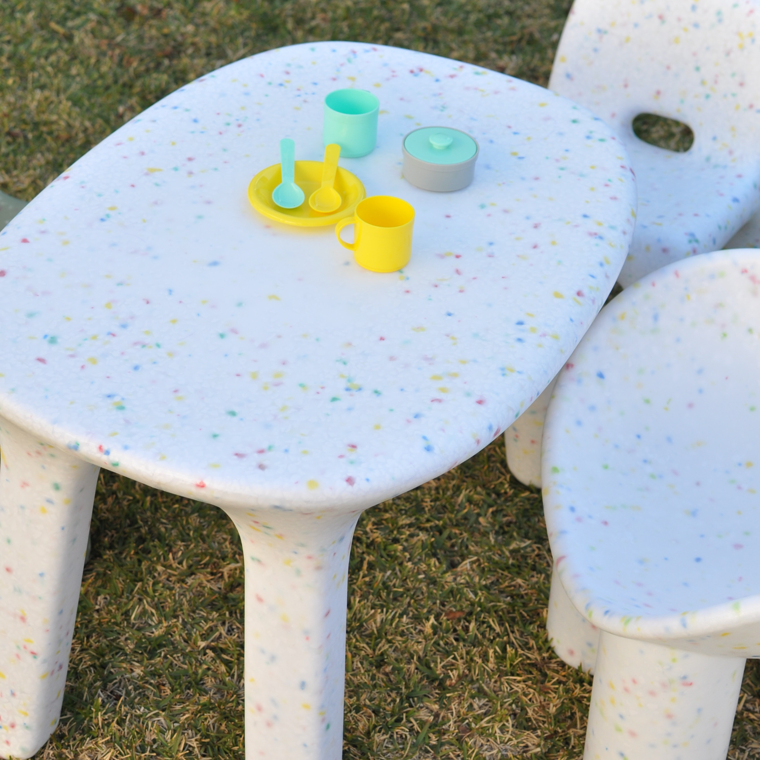 little boy sitting on white childrens speckles confetti table and chairs set with a red car on the table and grass background