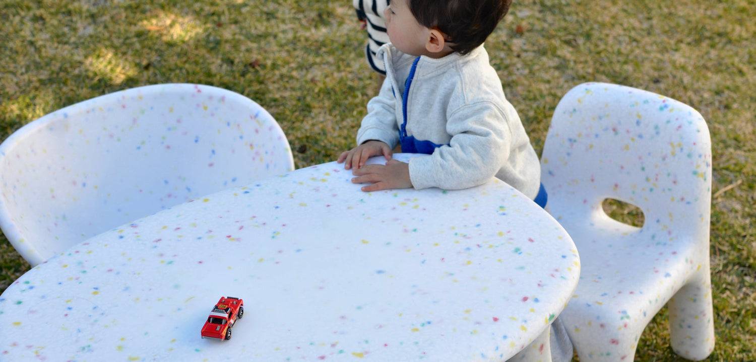 little boy sitting on white childrens speckles confetti table and chairs set with a red car on the table and grass background