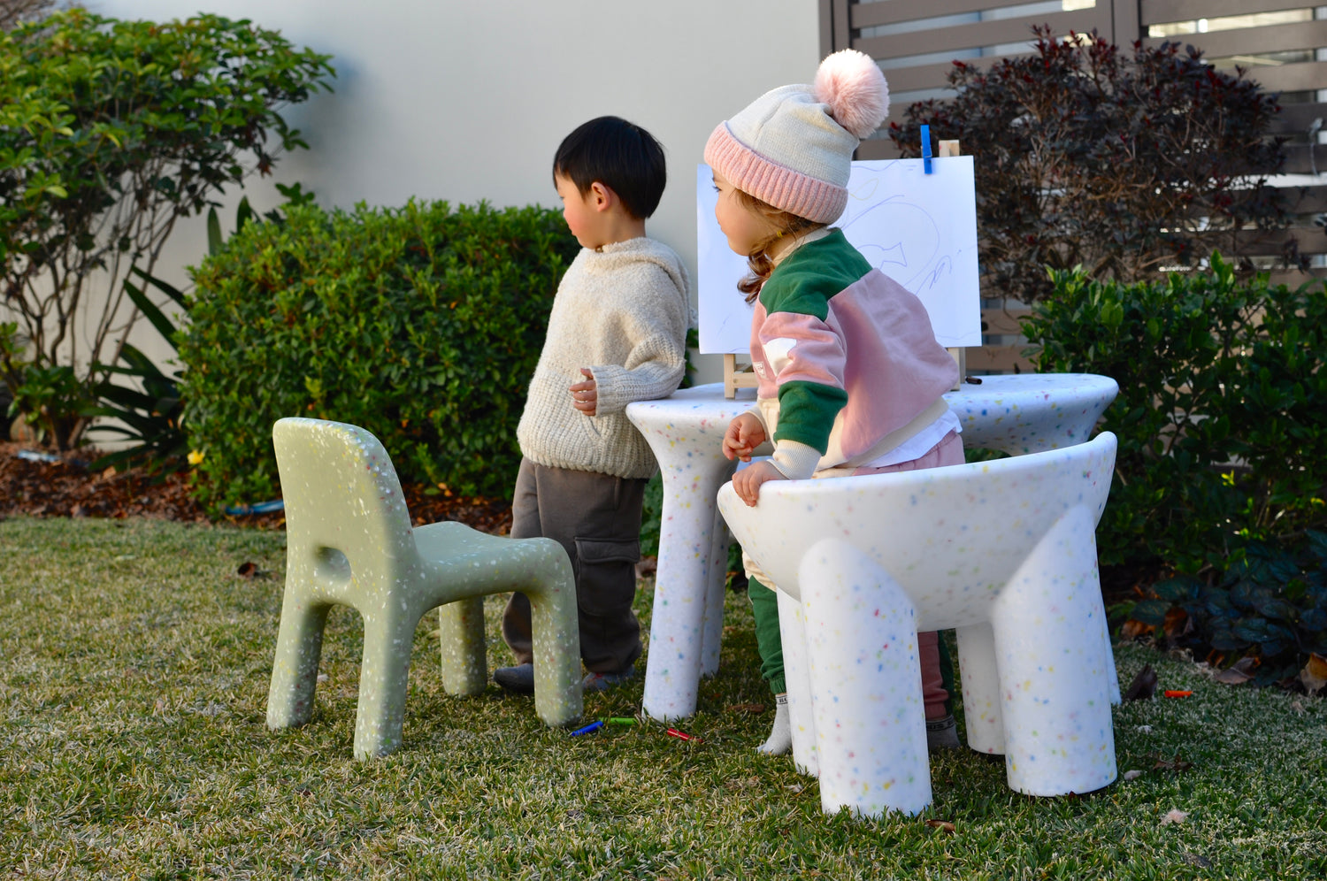 Two children sitting at a small speckles  confetti table and chair set outdoors with lots of greenery in the background. A photo shot to feature speckled children's outdoor furniture set