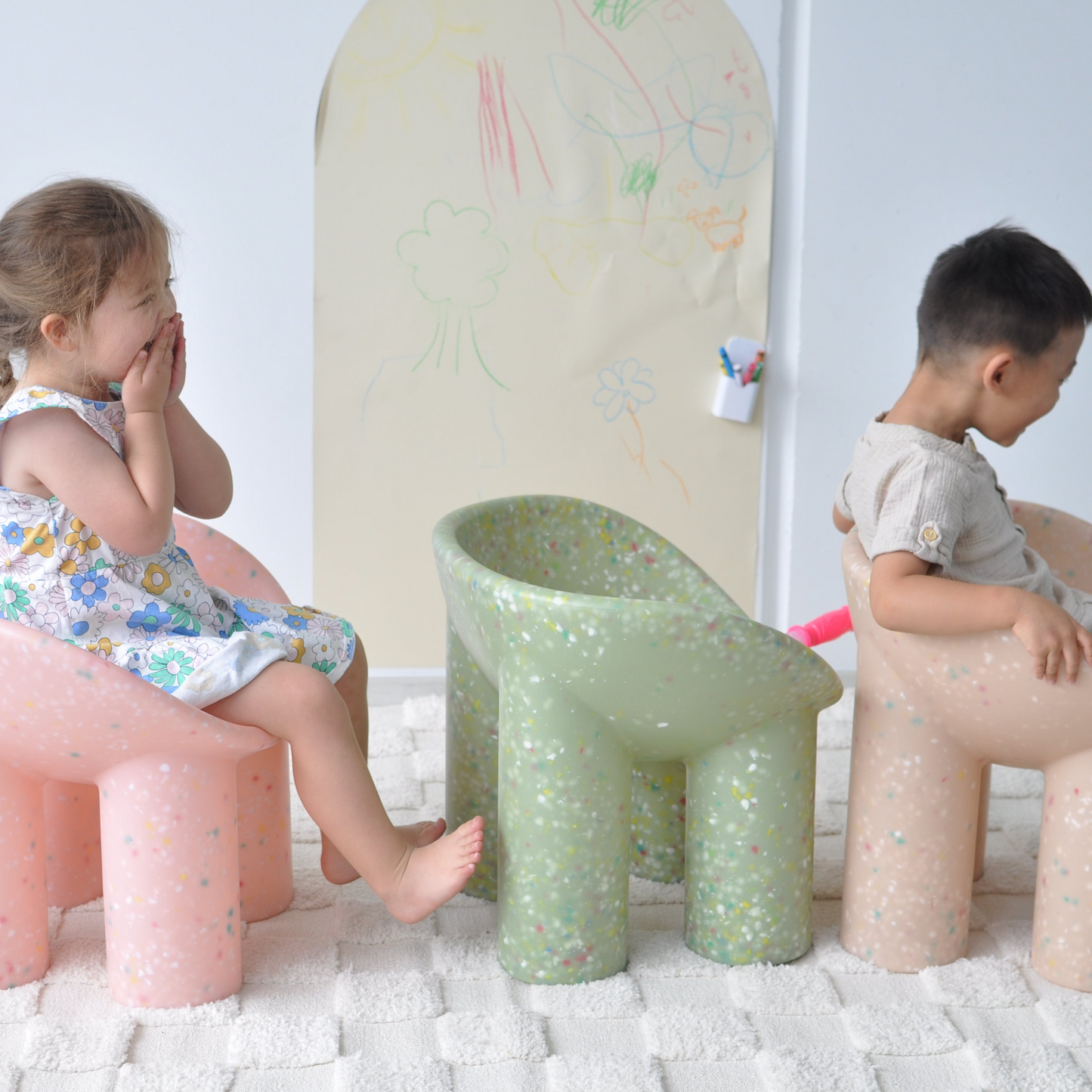 Two children sitting on colorful roly poly kids chairs in a room with a white wall and a child's drawing board