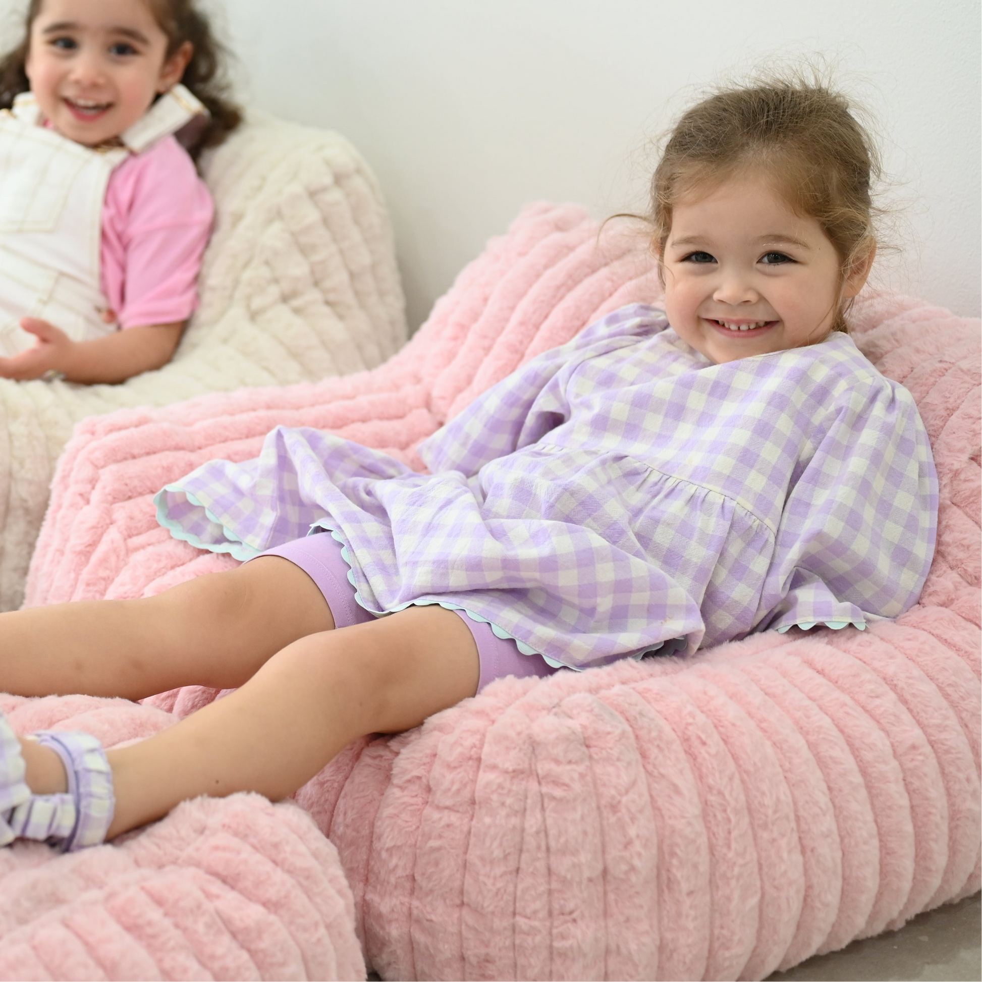 Two children in matching purple checkered outfits sitting on a pink bean bag chair.