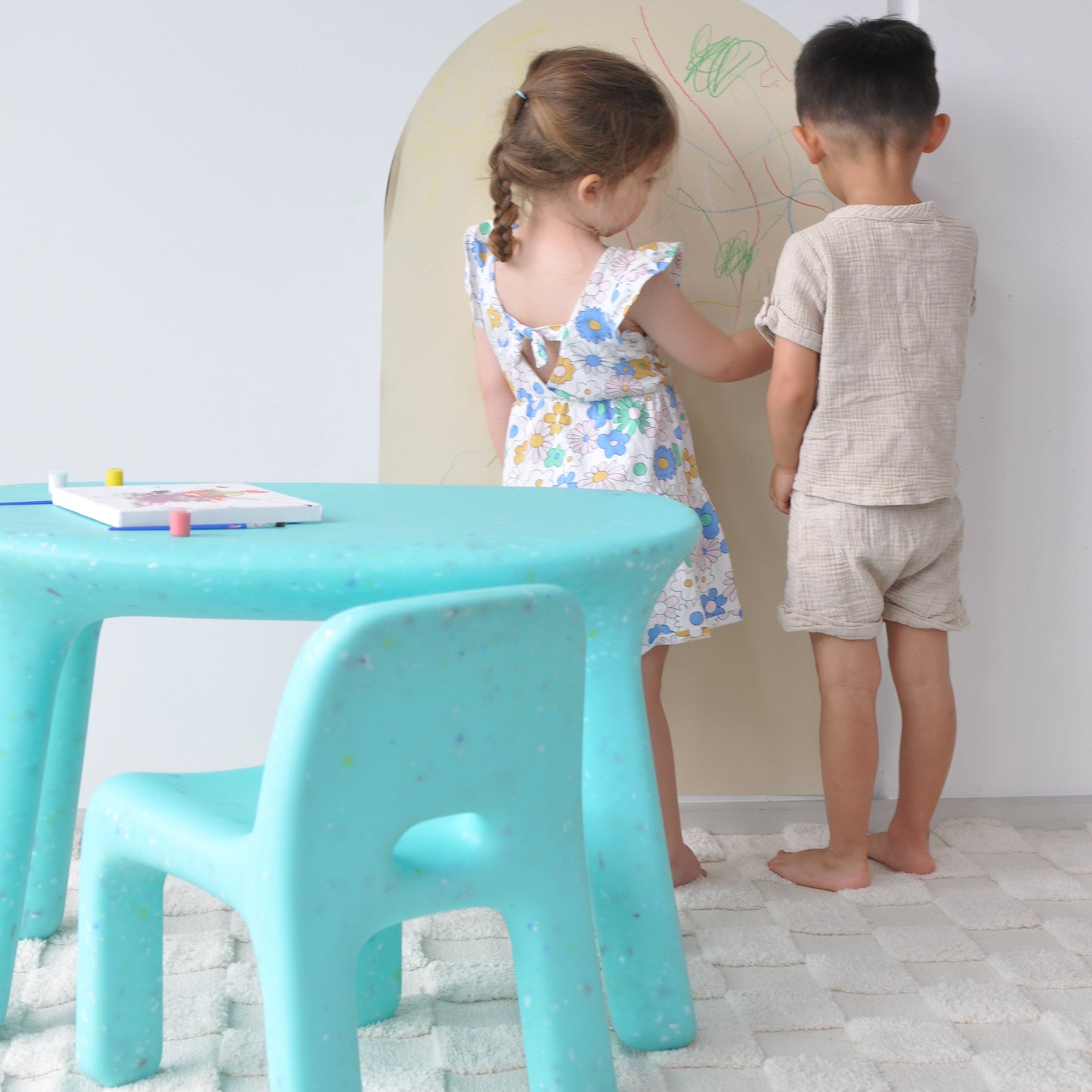 Two children playing with a green blue chair in a room with white walls.