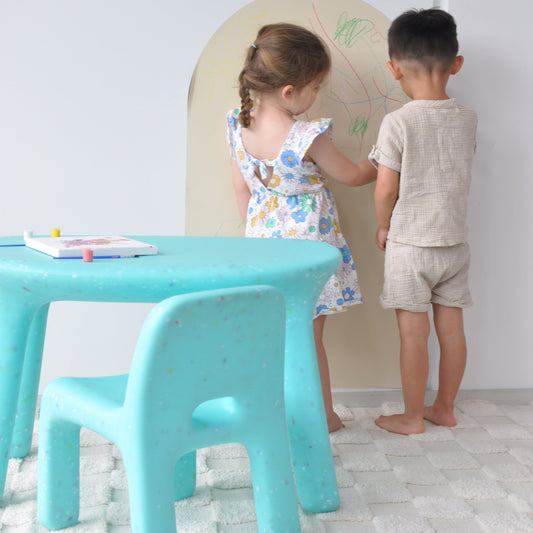Two children playing with a green blue chair in a room with white walls.