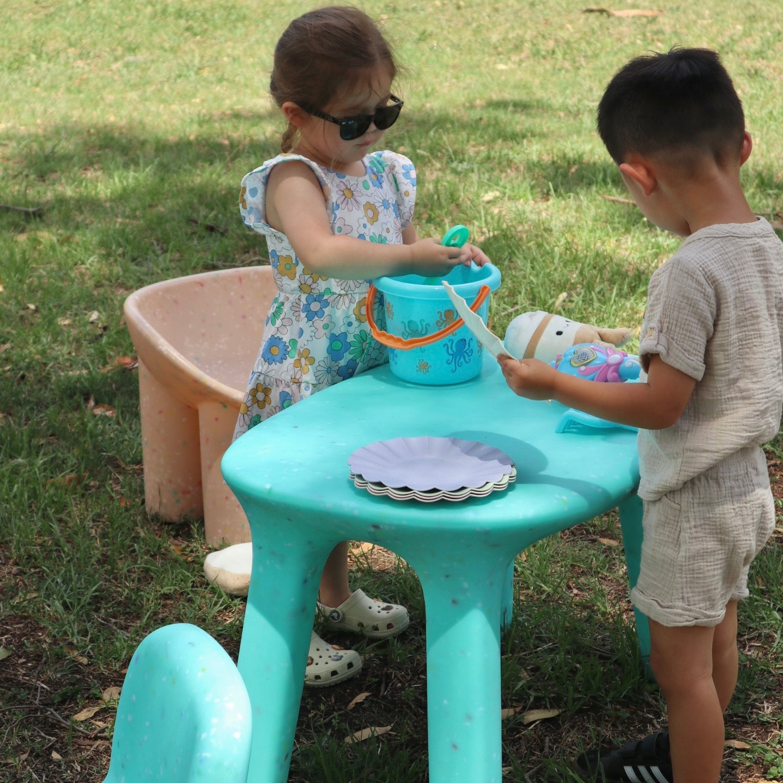 Two children playing on a confetti speckles kids turquoise table and beige roly poly chair on a grassy area.